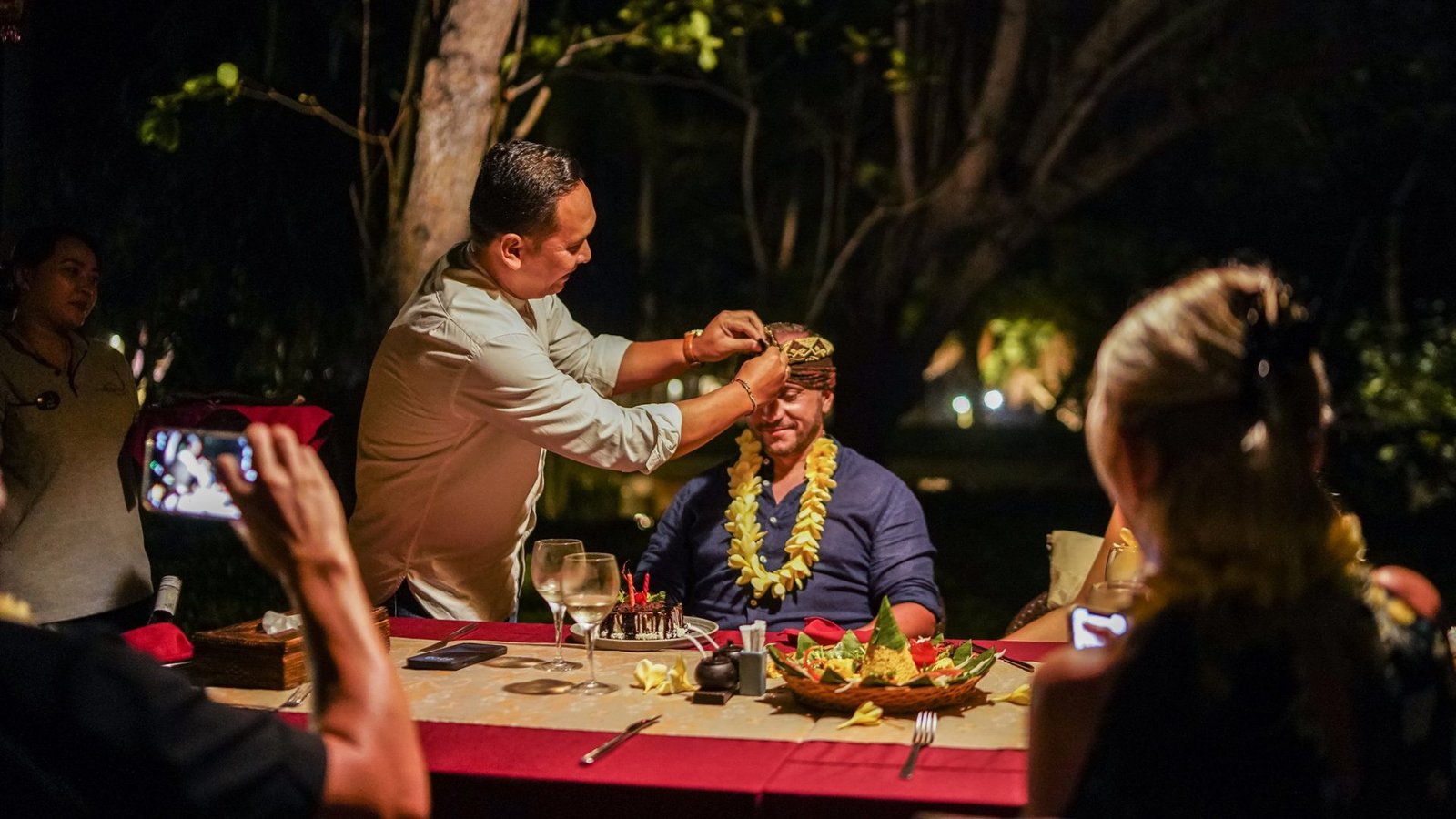 A host tying an udeng for a guest at a garden dinner